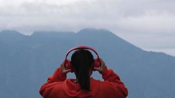 vista traseira de uma jovem de pé no topo de uma colina calma e ouvindo música em fones de ouvido pela manhã. mulher vestindo um suéter apreciando a beleza da natureza olhando para a montanha no inverno. video