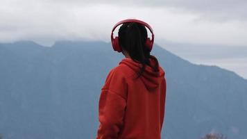 achterzijde visie van een jong vrouw staand Aan een kalmte heuveltop en luisteren naar muziek- in hoofdtelefoons in de ochtend. vrouw vervelend een trui genieten van de schoonheid van natuur op zoek Bij de berg in winter. video