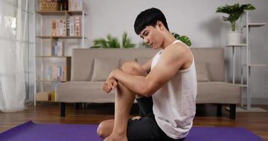 joven asiático sentado en el suelo y estirándose en la sala de estar en casa. ejercicio masculino en el piso interior por la mañana en casa. concepto de salud, ejercicio, fitness y estilo de vida. video