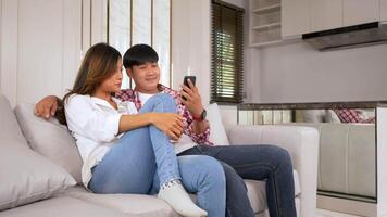 toma en cámara lenta, feliz pareja joven sentada en un sofá en la sala de estar en casa usa un teléfono inteligente charlando con un amigo, hablando y riendo juntos con felicidad, concepto de familia feliz video