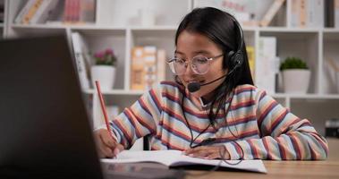 primer plano de gafas de niña con auriculares aprendiendo en línea a través de una computadora portátil mientras está sentado en el escritorio en casa. mujer joven escribiendo en el libro. concepto de educación y aprendizaje electrónico. video