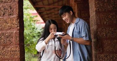 Happy Asian couple standing and looking photo on camera in ancient temple. Young man and woman selecting photo. Holiday, Lifestyle, travel and hobby concept.