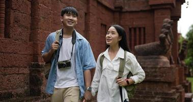 Happy asian traveler couple with hat hand together while visiting at ancient temple. Smiling young man and woman walking and looking ancient temple. Holiday, travel and hobby concept.