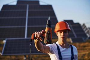 Cordless screwdriver in hand. Male worker in blue uniform outdoors with solar batteries at sunny day photo