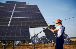 Using cordless screwdriver. Male worker in blue uniform outdoors with solar batteries at sunny day photo