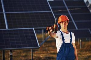 Cordless screwdriver in hand. Male worker in blue uniform outdoors with solar batteries at sunny day photo