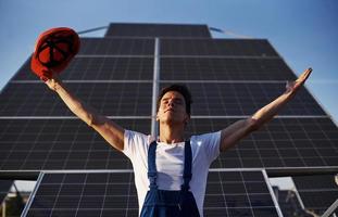 Hands raised up. Male worker in blue uniform outdoors with solar batteries at sunny day photo