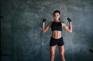 Young fitness woman is in the gym near wall with dumbbells in hands photo