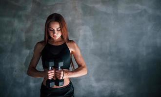 Young fitness woman is in the gym near wall with dumbbells in hands photo