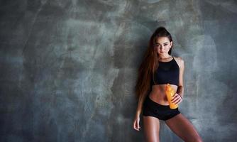 Young fitness woman with slim type of body and bottle of water is leaning on the wall photo