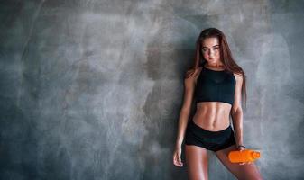 Young fitness woman with slim type of body and bottle of water is leaning on the wall photo