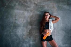 Young fitness woman with slim type of body and bottle of water is leaning on the wall photo