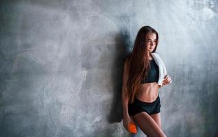 una joven mujer fitness con un tipo delgado de cuerpo y una botella de agua se apoya en la pared foto