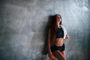una joven mujer fitness con un tipo delgado de cuerpo y una botella de agua se apoya en la pared foto