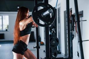 Leaning on the barbell. Young fitness woman with slim type of body and in black sportive clothes is in the gym photo