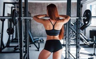 Leaning on the barbell. Young fitness woman with slim type of body and in black sportive clothes is in the gym photo