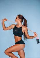 Stands with jumping rope. Young woman with slim body type isolated against blue background photo