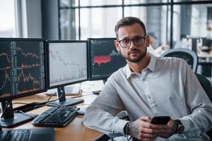 With phone in hands. Male stockbroker in formal clothes works in the office with financial market photo