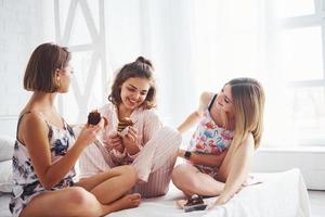 Eating sweets together. Happy female friends having good time at pajama party in the bedroom photo