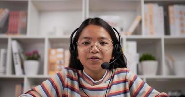 Headshot portrait of Asian girl glasses headphone talking and looking at camera while sitting at desk at home. Young female studying online via laptop and conferencing, discussing with teacher. video