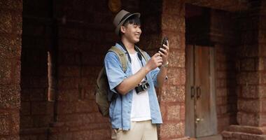 feliz viajante asiático com chapéu tira uma foto no smartphone e visita o antigo templo. homem jovem sorridente em pé e verificando no celular. conceito de férias, viagens e hobby. video