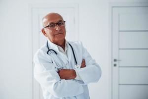 retrato de médico senior en uniforme blanco que se encuentra en la clínica foto