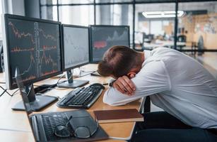 Tired businessman sleeping on the workplace by leaning on the table with multiple screens on it. Stock information on displays photo