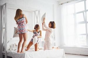 Pillow fight. Happy female friends having good time at pajama party in the bedroom photo