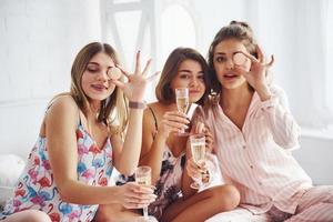 Celebrating with glasses of alcohol in hands. Happy female friends having good time at pajama party in the bedroom photo