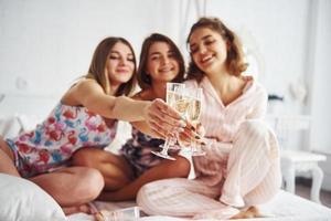 Celebrating with glasses of alcohol in hands. Happy female friends having good time at pajama party in the bedroom photo