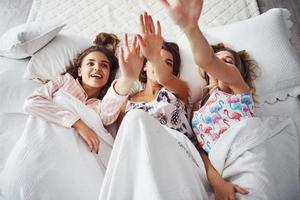 Lying on the bed under the blanket together. Happy female friends having good time at pajama party in the bedroom photo