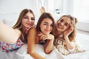 Making selfie. Happy female friends having good time at pajama party in the bedroom photo