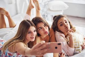 Making selfie by using pink phone. Happy female friends having good time at pajama party in the bedroom photo