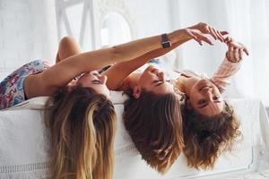 Lying on the bed with hair hangs down. Happy female friends having good time at pajama party in the bedroom photo