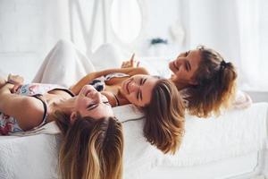 Lying on the bed with hair hangs down. Happy female friends having good time at pajama party in the bedroom photo