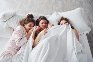 Hiding under the blanket. Happy female friends having good time at pajama party in the bedroom photo