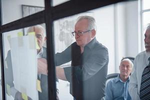 Working with stickers on glass. Aged team of elderly businessman architects have a meeting in the office photo