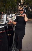 Blonde woman in sunglasses and in black dress near old vintage classic car photo