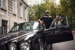 Blonde woman in sunglasses and in black dress near old vintage classic car photo