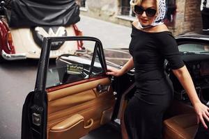 mujer rubia con gafas de sol y vestido negro saliendo del viejo coche clásico vintage foto