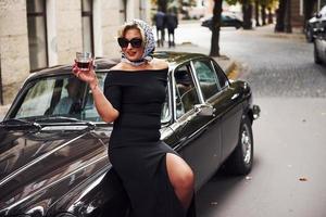 Blonde woman in sunglasses and in black dress near old vintage classic car photo