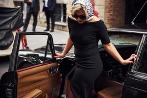 Blonde woman in sunglasses and in black dress going out from old vintage classic car photo