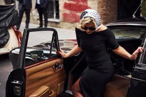 mujer rubia con gafas de sol y vestido negro saliendo del viejo coche clásico vintage foto