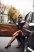 Blonde woman in sunglasses and in black dress nsits in old vintage classic car photo