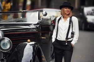 Blonde woman in hat and in black retro clothes near old vintage classic car photo