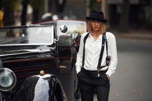 mujer rubia con sombrero y ropa retro negra cerca del viejo coche clásico vintage foto