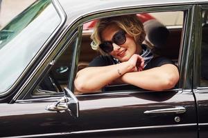 mujer rubia con gafas de sol y vestido negro se sienta en un viejo coche clásico vintage foto