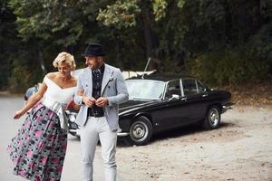 Beautiful couple in old fashioned wear near retro car with forest at background photo