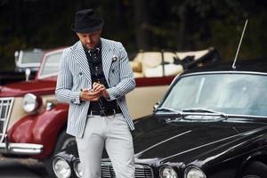 Man in formal clothes and black hat stands near retro automobiles photo