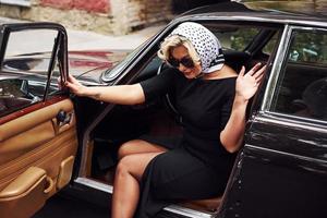 mujer rubia con gafas de sol y vestido negro saliendo del viejo coche clásico vintage foto